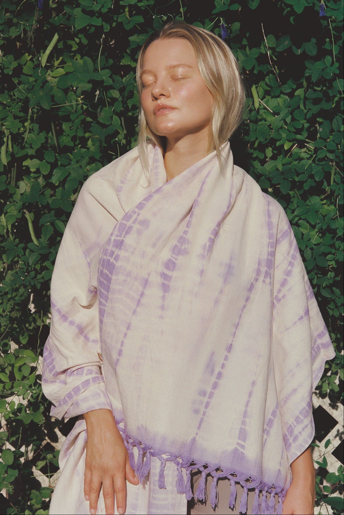 Woman wearing a purple tie-dye yoga towel with purple accents against a green leafy background